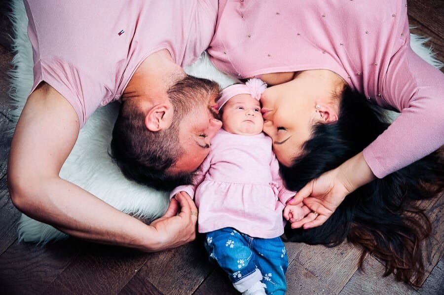 BABY- und Familienfotografie: Eltern und kleines Mädchen liegen auf dem Boden, Kopf an Kopf, und schauen liebevoll auf das Baby, das in der Mitte liegt, in einem hellen Raum.