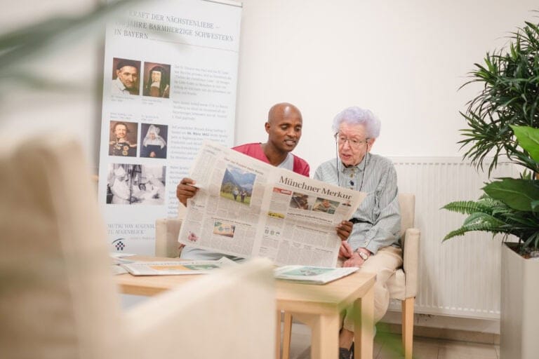 Menschen beim gemeinsamen Lesen einer Zeitung in einem Wartezimmer mit Pflanzen im Hintergrund.