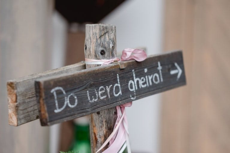 Schwarz-weißer Holzschild mit "Do werd gheirat" und Pfeil, dekoriert mit rosa Schleifen, für Hochzeiten oder Abschiede.