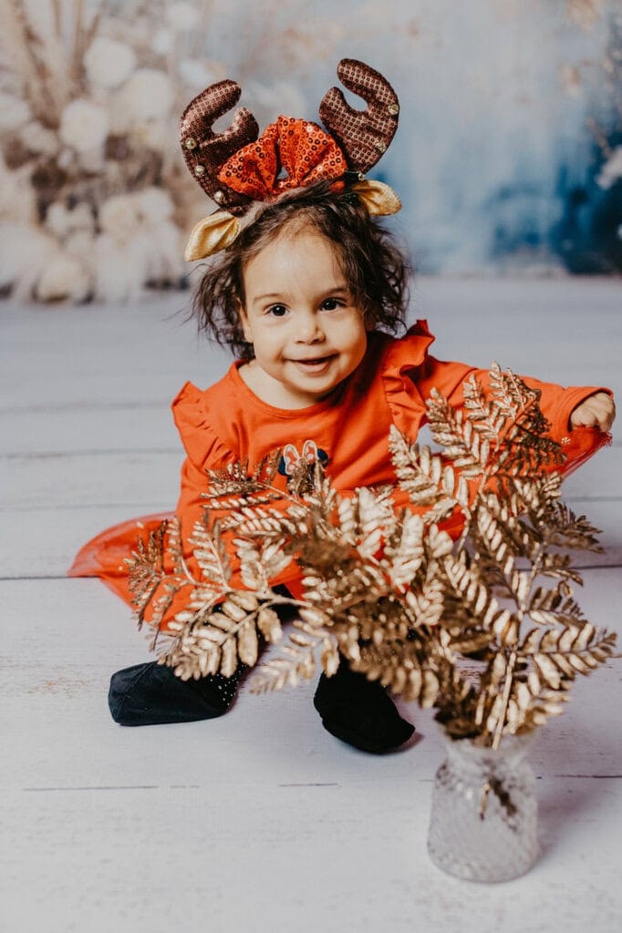 Fröhliches Kind in festlichem Weihnachten-Outfit mit Rentier-Haarreif, auf Holzboden sitzend, schön dekorierter Hintergrund, professionelle Fotografie, Kinderfotostudio München, festliche Kinderfotos, Weihnachtsbilder, kindgerechte Porträts, Shooting für Familien und Kinder in München.