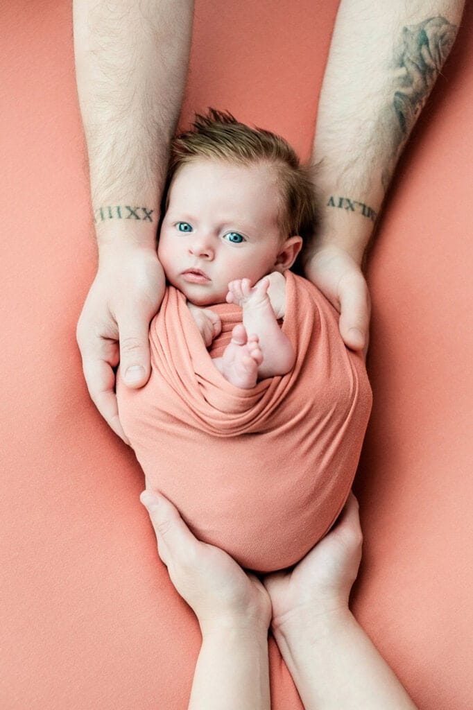 Father with tattooed arms holds his baby wrapped in a salmon-colored cloth.