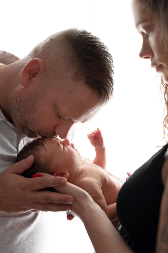 Father tenderly kisses his baby's forehead in bright backlighting.