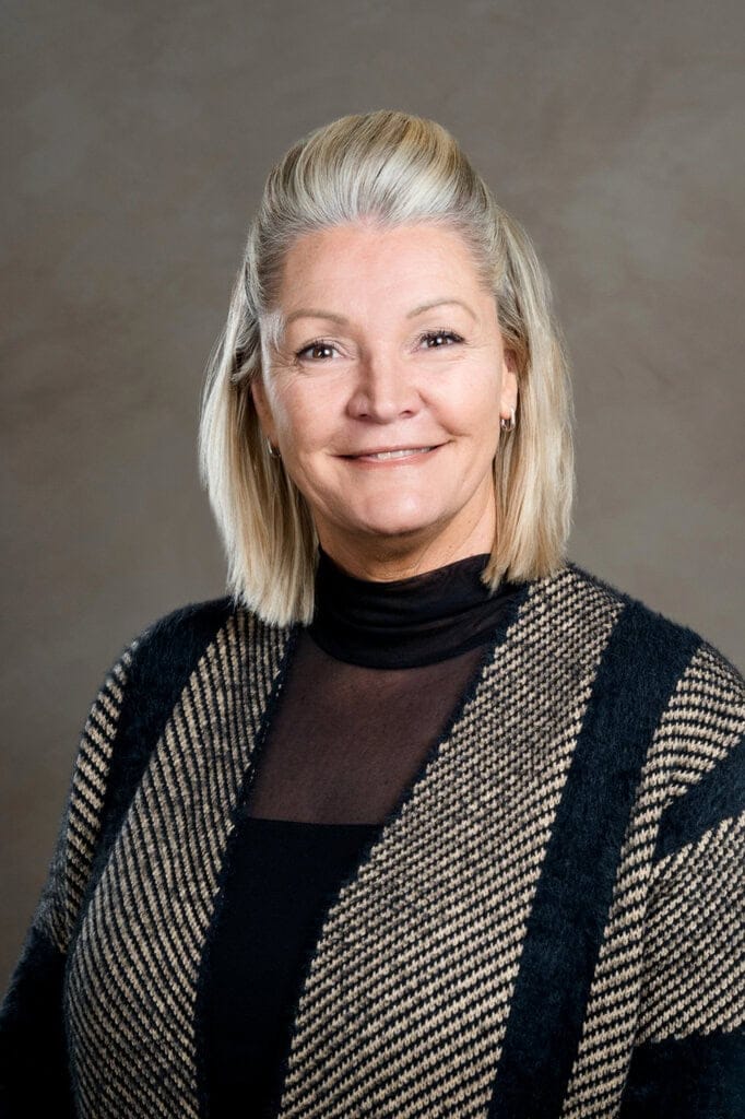 Freundliche Frau mit blonden Haaren in gemusterter Strickjacke.