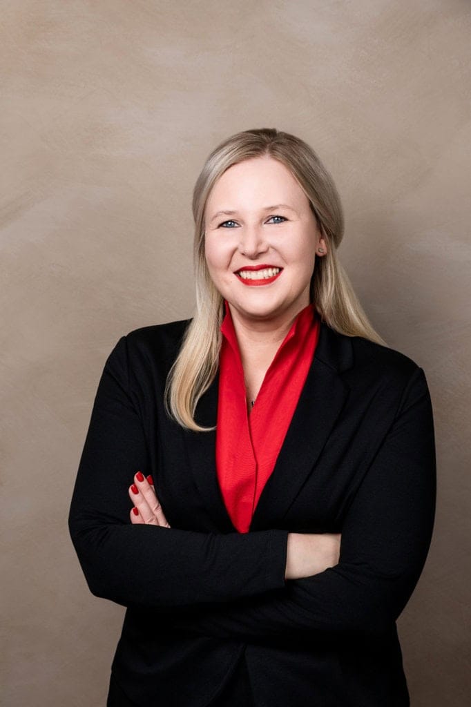 Attraktives Businessporträt einer professionellen Frau in schwarzem Blazer mit rotem Schal, pose mit verschränkten Armen vor neutralem Hintergrund, ideale Vorlage für Business- und Bewerbungsfotos.