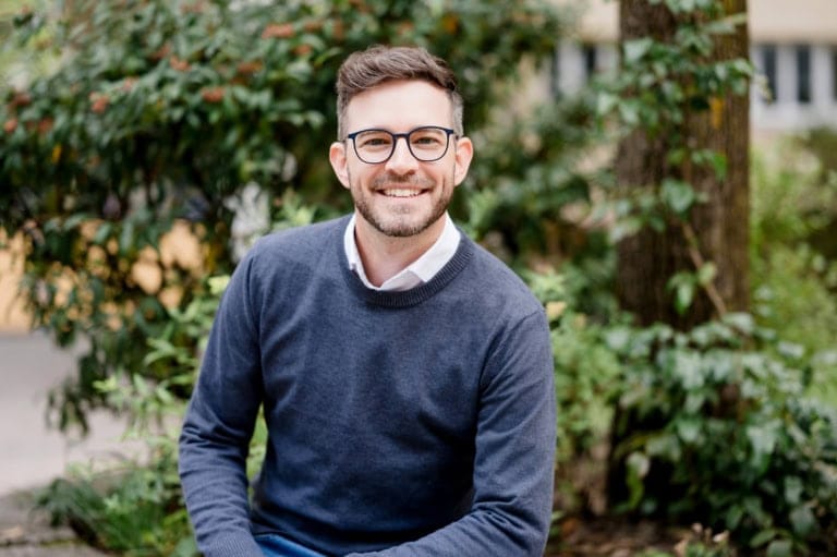 Sympathischer Mann mit Brille und blauem Pullover im Grünen fotografiert.