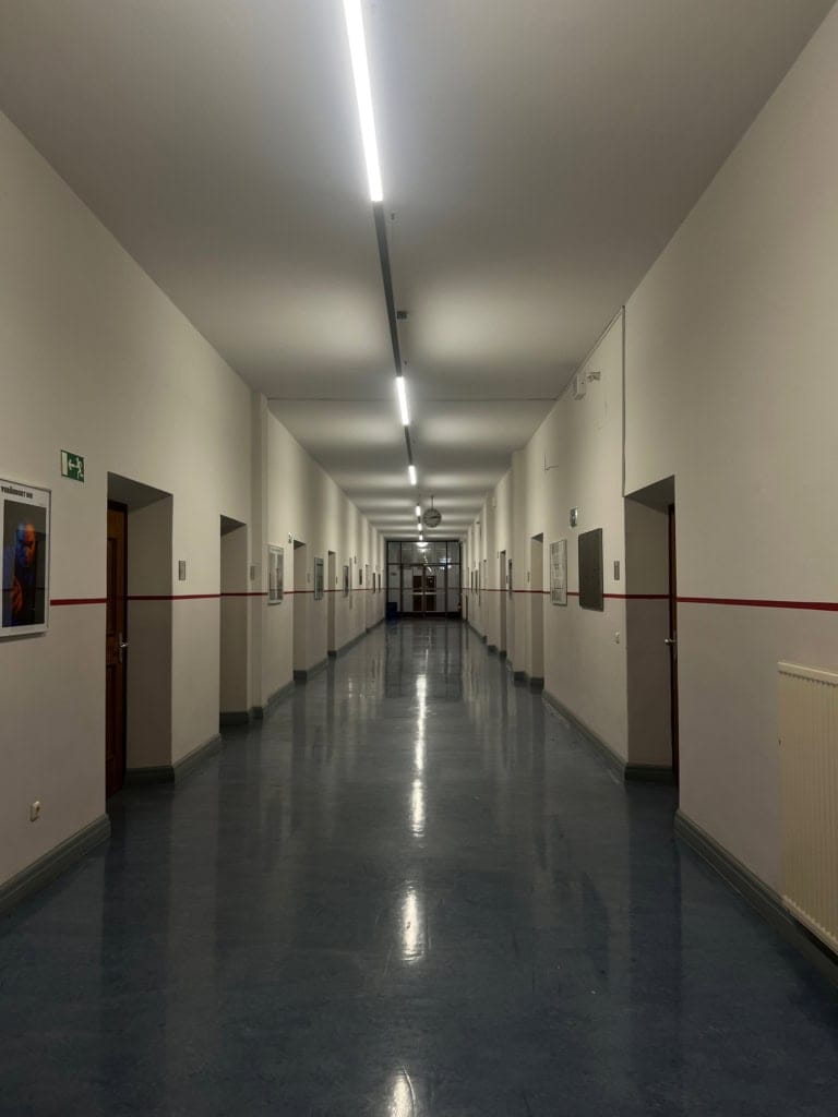 Leere Flur in einem Schulgebäude, moderne Beleuchtung, helle Wände, Zeitungs- oder Posterrahmen, ruhiges Ambiente, klare Linien, reflektierende Fußböden, ruhige Atmosphäre, professionell aufgenommen.
