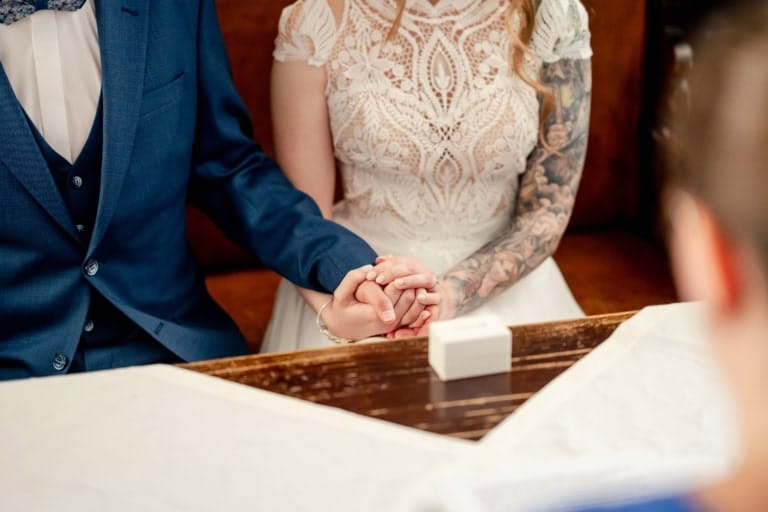 Nahaufnahme eines Hochzeitspaares bei der Trauung, das sich die Hände hält, mit Fokus auf die Hände und das Brautkleid.