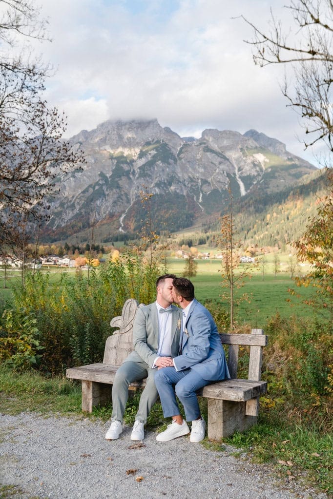 Romantischer Kuss zweier Bräutigame auf einer Holzbank mit Bergblick.