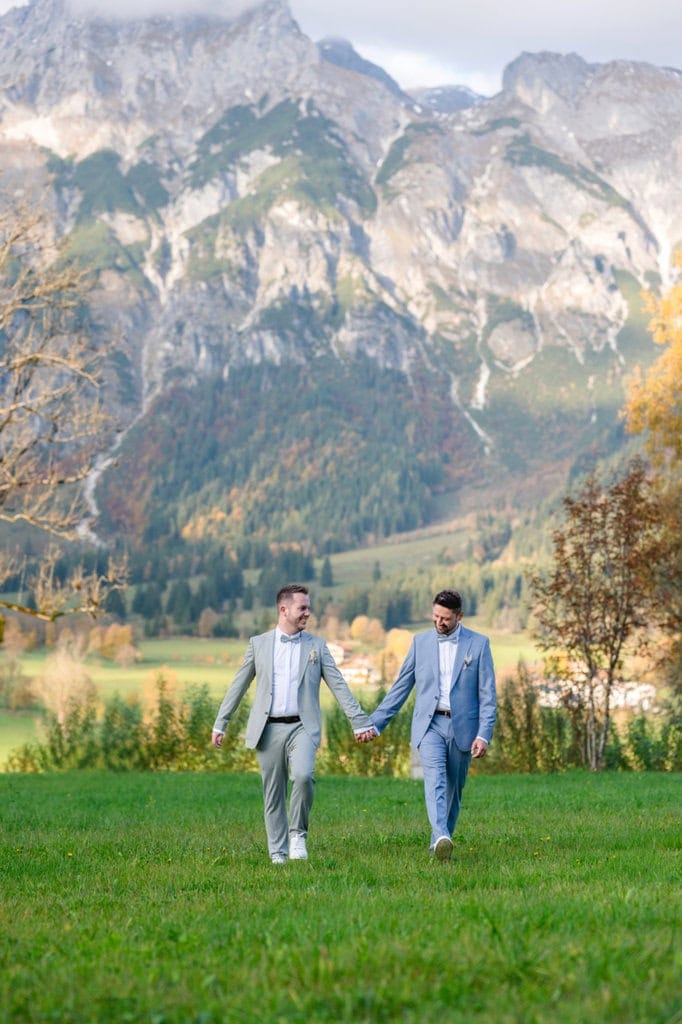 Männer-Hochzeitspaar spaziert Hand in Hand über eine grüne Wiese.
