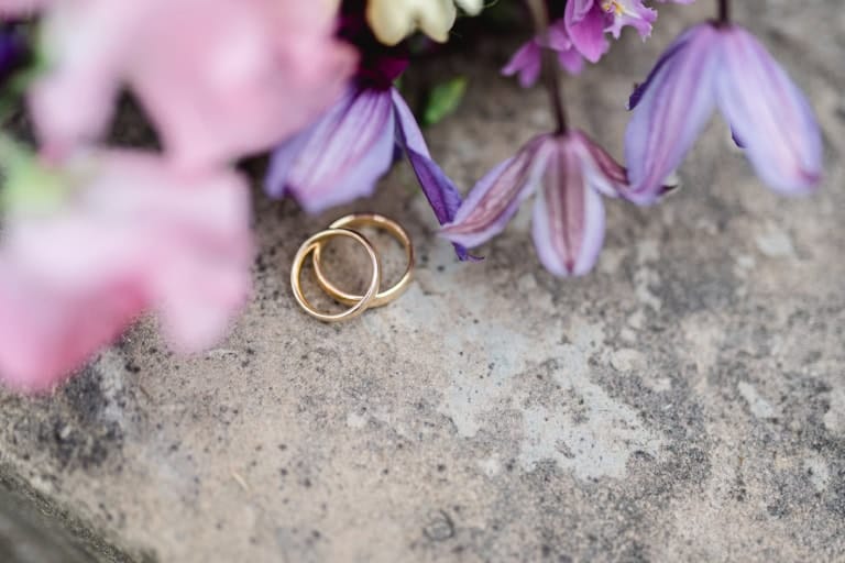 Elegante Detailaufnahme der goldenen Eheringe auf Stein mit lila Blüten.